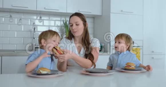 年轻的家庭妈妈和两个儿子坐在厨房的白色桌子上一起吃汉堡玩得很开心儿童视频的预览图