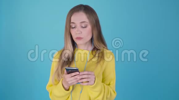 漂亮的金发女郎穿着黄色毛衣听着耳机里的音乐在蓝色背景上唱歌可爱的女孩视频的预览图
