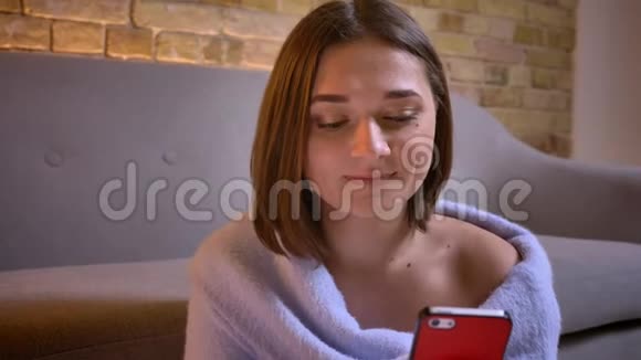 一位年轻迷人的白种人女性用手机看着摄像机在舒适的室内家庭微笑特写镜头视频的预览图