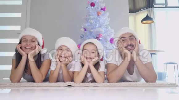 滑稽的爸爸妈妈和戴着红袜子和帽子的孩子们躺在圣诞树附近的地毯上双手紧握着视频的预览图
