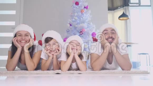 滑稽的爸爸妈妈和戴着红袜子和帽子的孩子们躺在圣诞树附近的地毯上双手紧握着视频的预览图