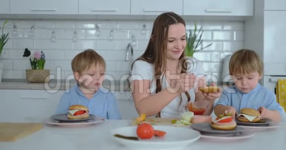 年轻漂亮的妈妈穿着白色连衣裙带着两个孩子微笑着在厨房里吃着新鲜的汉堡视频的预览图