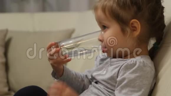 小孩子坐在房间的沙发上喝着玻璃杯里的水视频的预览图