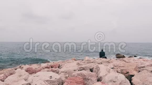 孤独悲伤的人坐在靠近海洋的大石头上上班族每天下班后坐在海边孤独的男孩望着大海视频的预览图