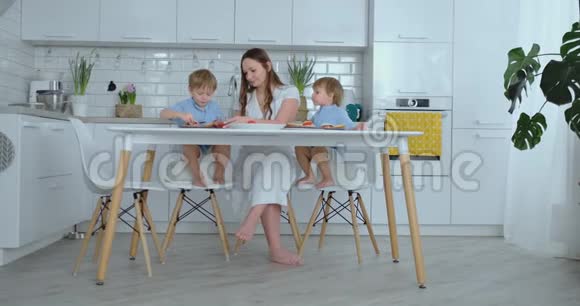 年轻漂亮的妈妈穿着白色连衣裙带着两个孩子微笑着在厨房里吃着新鲜的汉堡视频的预览图