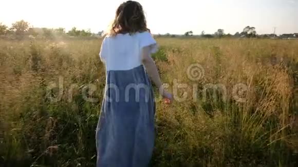 穿着浅色连衣裙的女人在日落时穿着高草穿过田野动作缓慢镜头移动视频的预览图