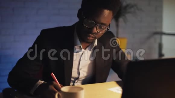 年轻的黑人商人带着笔记本电脑和文件在夜间办公室工作他在文件上签字喝咖啡视频的预览图