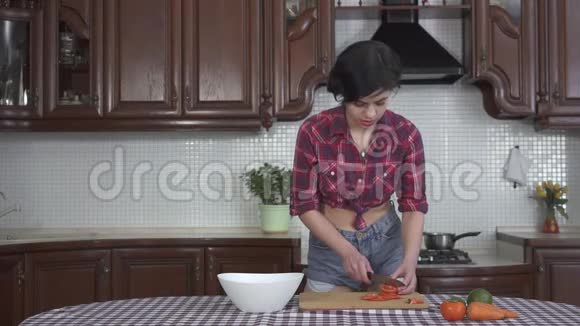 穿着格子衬衫和短裤穿着现代舒适的厨房里的木板上切菜的漂亮微笑的年轻女孩视频的预览图
