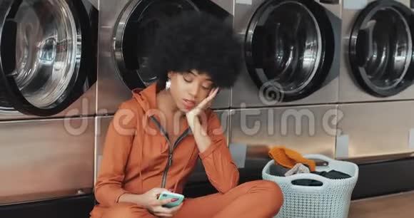 疲惫的年轻非洲裔美国妇女在自助洗衣店使用智能手机她等待这个过程的完成视频的预览图