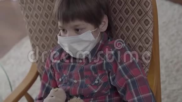 肖像小生病的男孩坐在扶手椅上脸上戴着医疗面具拿着玩具在家里不同的药丸视频的预览图