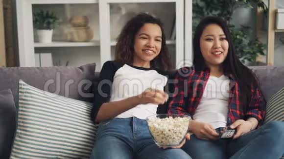 非裔美国人和亚裔女青年在家里享受电视上的喜剧笑说吃爆米花放松身心视频的预览图