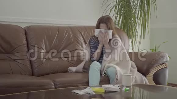 十几岁的女孩裹着毯子坐在家里的皮革沙发上用餐巾纸吹着鼻子女孩视频的预览图