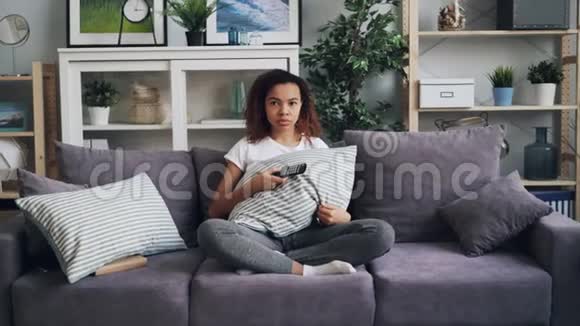 一位年轻的非洲裔美国女孩在电视上看恐怖电影躲在枕头后面坐在沙发上抱着视频的预览图
