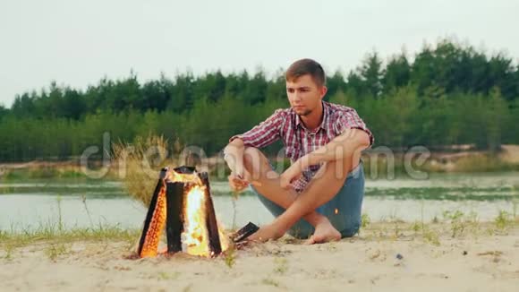 孤独严肃的男人坐在篝火旁用棍子烤棉花糖视频的预览图