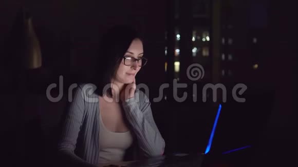 可爱的年轻女子在黑暗中微笑着看着家里的笔记本电脑屏幕视频的预览图