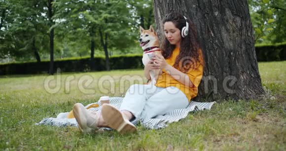 女人带着狗在绿色公园用智能手机耳机听音乐视频的预览图