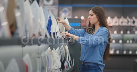 穿着蓝色衬衫和牛仔裤的漂亮黑发女人在消费电子商店里选择了熨斗视频的预览图