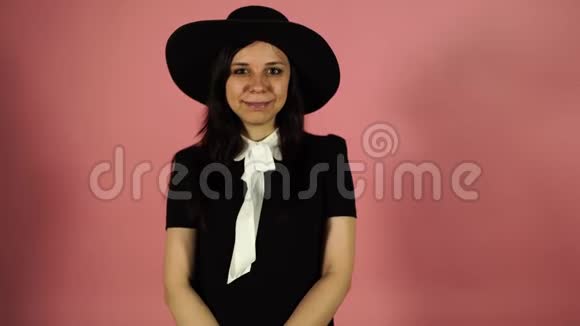 女士在粉红色的背景上微笑穿黑色连衣裙和帽子的女人女性眨眼概念调情视频的预览图