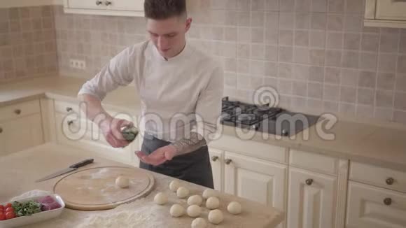 年轻的比萨饼师穿着厨师制服把橄榄油倒在手上在厨房里擀面团做比萨饼食品准备视频的预览图
