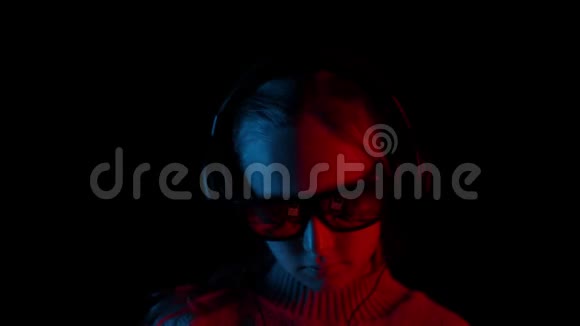年轻女孩戴着耳机听音乐在黑暗的工作室与红色和蓝色照明喜欢移动音乐的少女视频的预览图