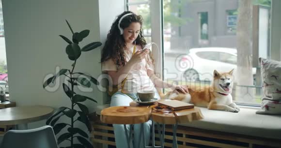 女孩用耳机听音乐用智能手机在咖啡馆和狗视频的预览图
