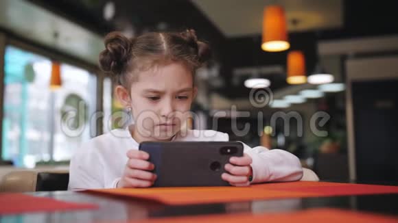 一个小女孩坐在咖啡馆的桌子旁喜欢在智能手机上玩游戏视频的预览图