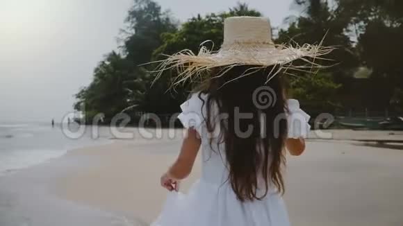 镜头跟随快乐无忧无虑美丽的女孩享受轻松的散步沿着大气的海滩戴着大草帽视频的预览图