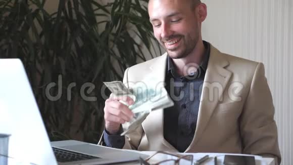 年轻的快乐商人穿着西装坐在他的工作场所手里拿着一捆钱成功的商业人士视频的预览图