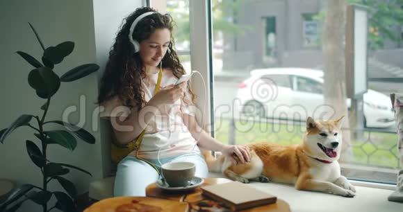 戴着耳机的女人在咖啡馆里用智能手机抚摸宠物听音乐视频的预览图