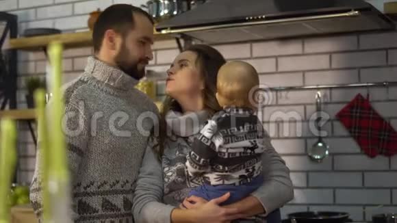 大胡子的父亲穿着温暖的毛衣在厨房里和抱着妈妈的小儿子玩耍女人给孩子视频的预览图