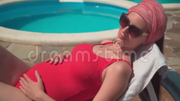 怀孕的女孩穿着红色的一件泳衣和眼镜躺在游泳池附近的休息室里抚摸着她的肚子视频的预览图