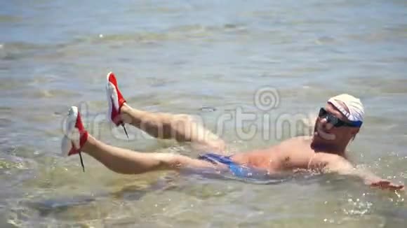 一个穿着泳裤戴着太阳镜穿着夹克的人躺在浅水里在阳光下溅起水花视频的预览图