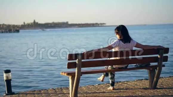 夏天夕阳西下海边筑堤一位年轻的黑发女子坐在长凳上背影欣赏着美丽的日落孤独寂寞视频的预览图