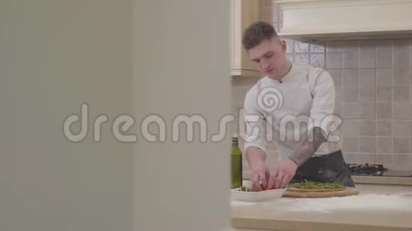 穿着厨师制服的年轻人把西红柿樱桃放在披萨的顶部躺在桌子上专业烹饪视频的预览图