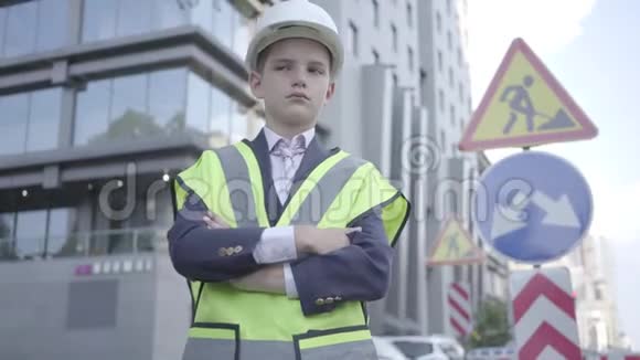 肖像可爱的小成功男孩穿着商务服安全设备和建筑工人头盔站在繁忙的地方视频的预览图
