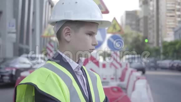 可爱的小成功男孩穿着商务服安全装备和建筑工人头盔站在繁忙的道路上视频的预览图