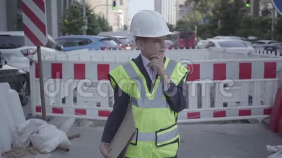 一个忧郁的小男孩戴着安全设备和建筑工人头盔的肖像手持建筑计划站在一个视频的预览图