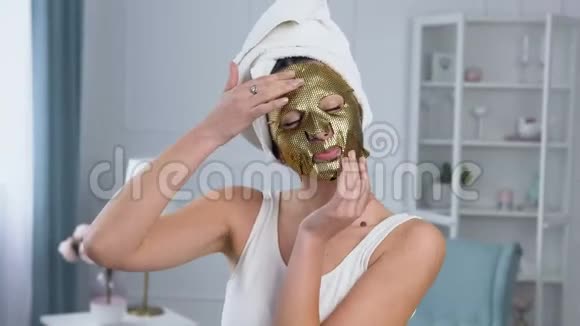 英俊的微笑女人在脸上固定恢复活力的化妆品金色组织面具的肖像视频的预览图