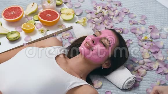 漂亮的年轻女人带着粉红色的面具在健康中心放松而美容师则把黄瓜放在眼睛上视频的预览图