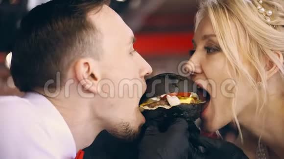 特写肖像新郎新娘吃一个大黑汉堡一个三明治玩得开心玩得开心看着视频的预览图