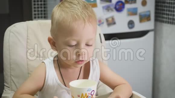 可爱的小男孩在家吃早餐时喝牛奶同时在平板电脑上看卡通儿童和技术视频的预览图
