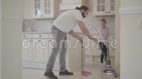 爸爸留着胡子小可爱的女儿用扫帚和簸箕在新的现代化厨房里打扫卫生视频的预览图