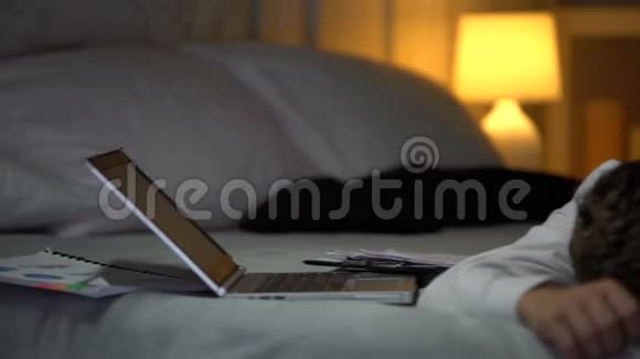 工作过度的商人在文件和笔记本电脑附近的床上睡着了烧光了视频的预览图
