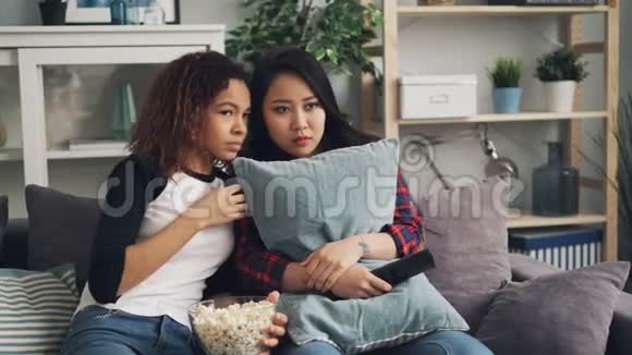 情绪激动的年轻女性正在一起看恐怖电影躲在枕头后面闭上眼睛女孩们在吃爆米花视频的预览图