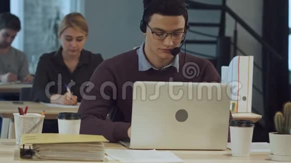 在电话中心工作的快乐的年轻工人带着耳机微笑在笔记本电脑上工作视频的预览图