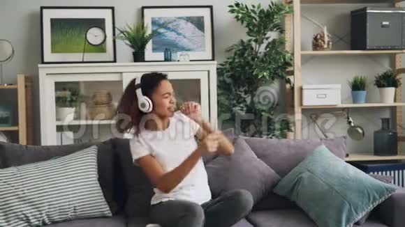 可爱的非裔美国女孩正在用耳机听音乐在现代的沙发上唱歌跳舞视频的预览图