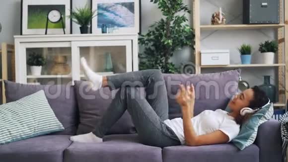 可爱的非裔美国女孩正在耳机里听音乐双手双腿躺在沙发上生活视频的预览图