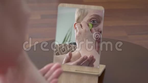漂亮的老妇人看着镜子在家里用一片黄瓜擦着脸化妆品视频的预览图