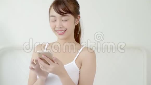 美丽的肖像年轻的亚洲女人微笑着坐着看着智能手机阅读社交网络与享受床上卧室视频的预览图