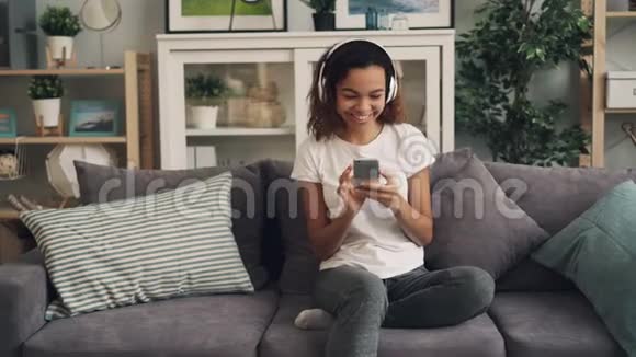 微笑非洲裔美国女孩正在通过耳机听音乐并使用智能手机在家里的沙发上放松视频的预览图